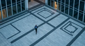 High aerial drone shot of a person walking on a geometrically patterned urban plaza at dusk, smartphone glowing with cool blue light, surrounded by modern buildings.