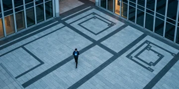 High aerial drone shot of a person walking on a geometrically patterned urban plaza at dusk, smartphone glowing with cool blue light, surrounded by modern buildings.