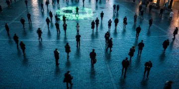 Bird's-eye view of a wet, neon-lit city square at dusk, showing blurred pedestrians engrossed in smartphones, reflecting the pervasive and integrated digital lifestyle.
